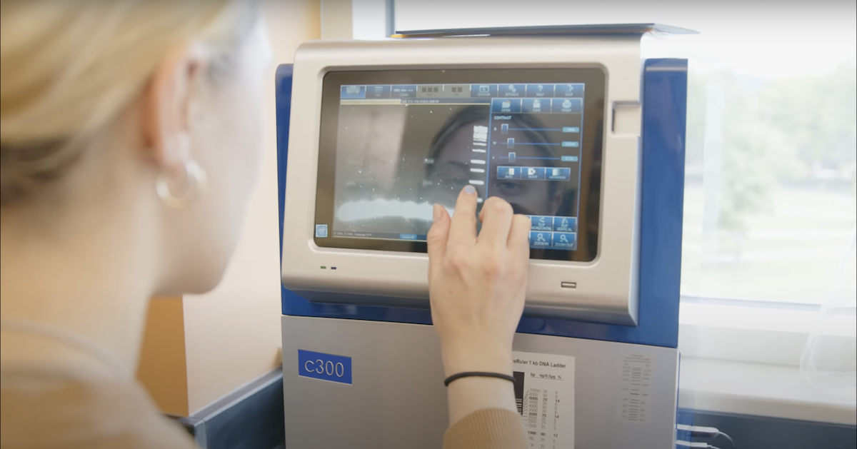 A person with light hair is using a touch screen on a laboratory c300 machine. The screen displays various technical options and settings. The person is interacting with the screen in a bright indoor setting.
